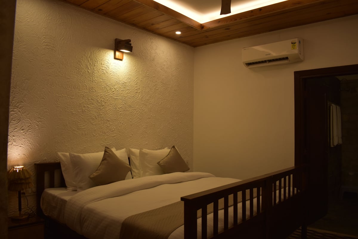 A bedroom with a bed, pillows, a nightstand, a lamp, a wooden ceiling, and an air conditioning unit.