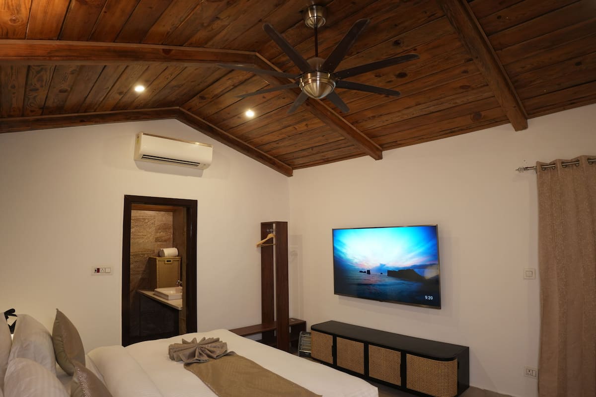 A bedroom with a wooden ceiling, a bed with pillows, a TV on the wall, and a small shelf with toiletries.