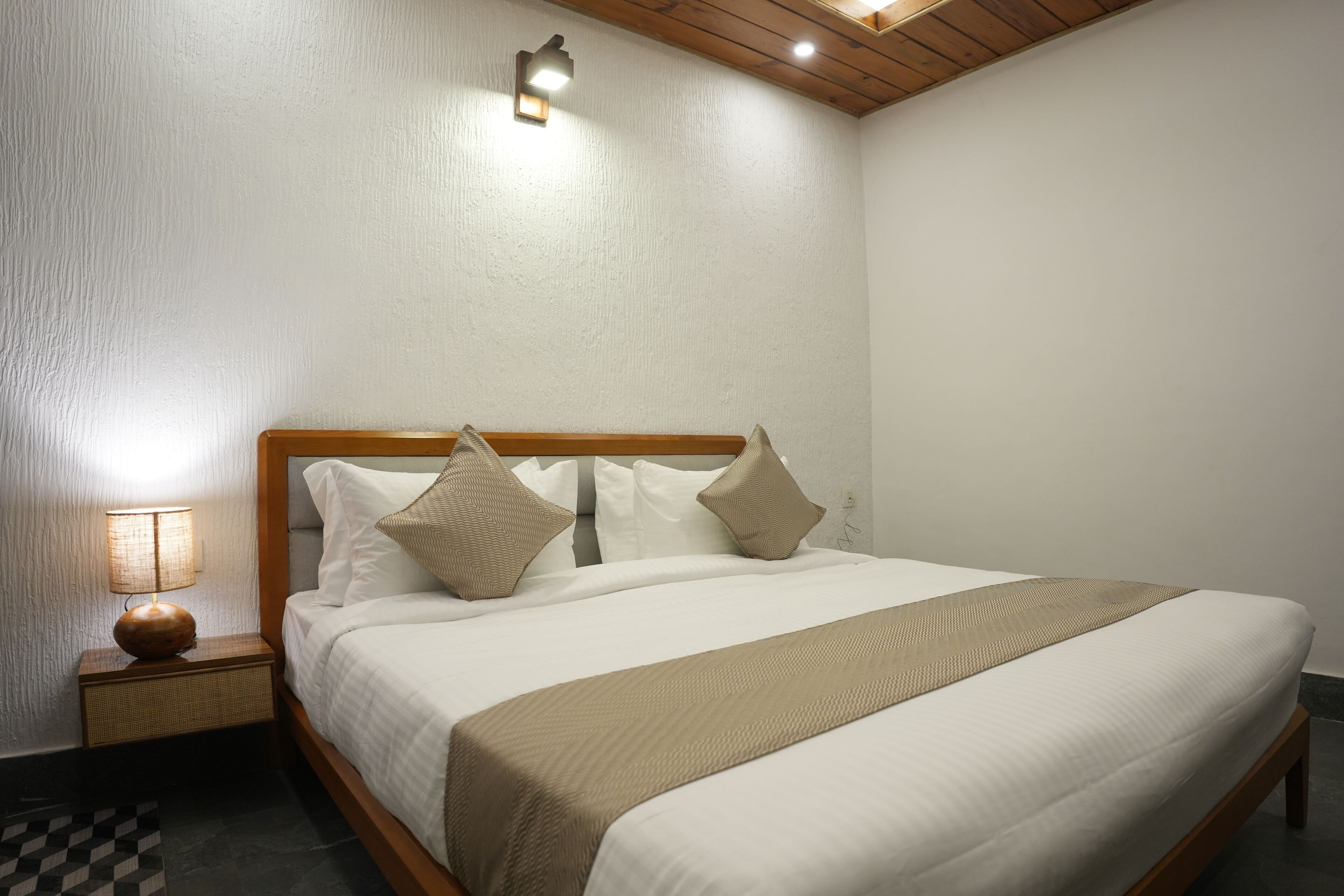A neatly made bed with white linens and two beige pillows, a wooden headboard, and a bedside lamp on a nightstand.