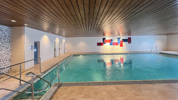 An indoor swimming pool with a wooden ceiling, tiled walls, and a clear blue pool.