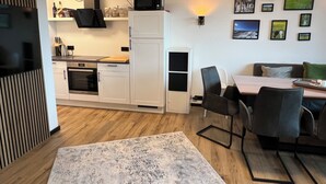 A modern kitchen with white cabinets, a microwave, and a coffee machine. There is a dining area with a table and chairs, and a sofa.