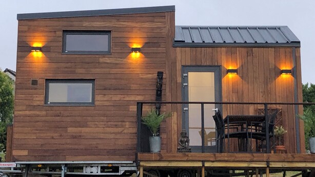 Een modern houten gebouw met twee balkons, waarvan er één een tafel en stoelen heeft, en twee verlichte ramen aan de zijkant.