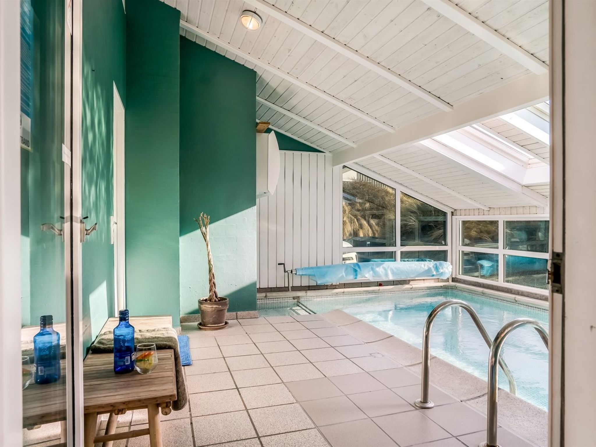 A pool area with a glass wall, a table with bottles, and a potted plant.