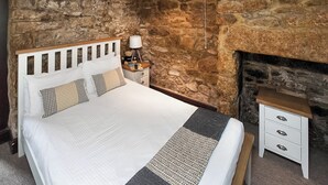 A bedroom with a stone wall, a bed with white bedding, a bedside table with a lamp, and a small wooden bench. - The Summerhouse 2 Bed (Penzance)