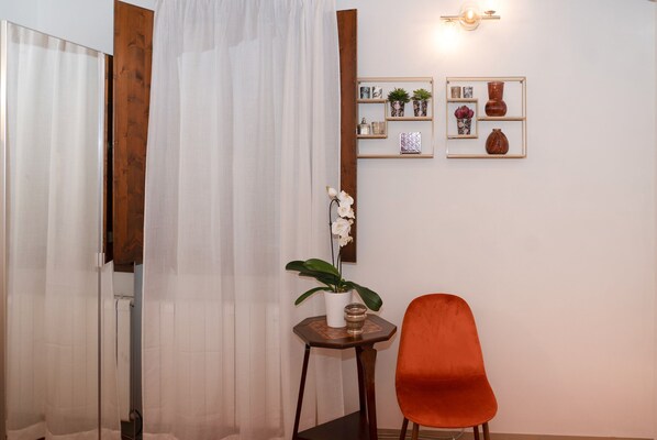 A room with a wooden door, a white curtain, a small table with a potted plant, an orange chair, and wall-mounted decor including a mirror and framed pictures.