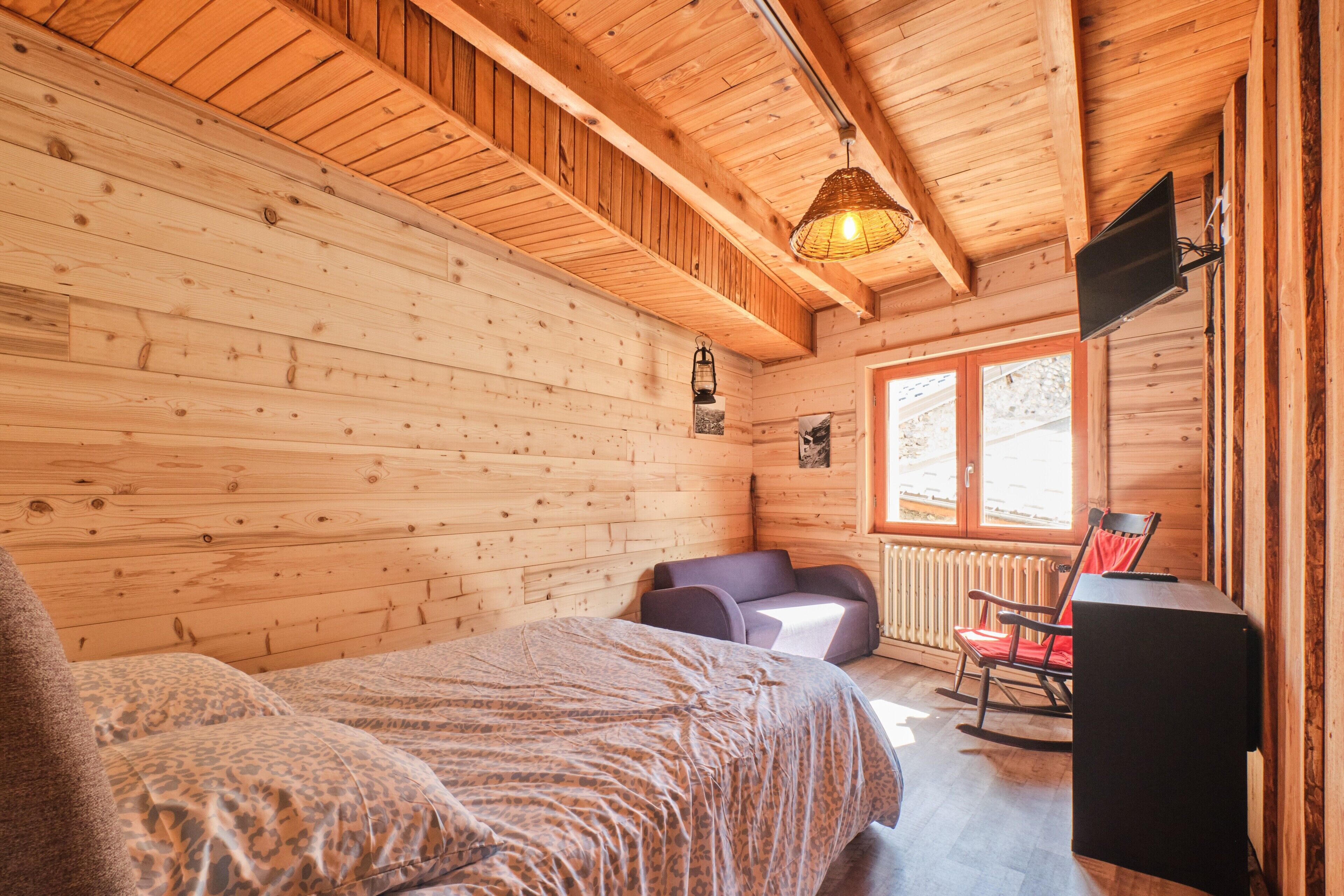 A wooden cabin room with a bed, a rocking chair, a television, and a window.