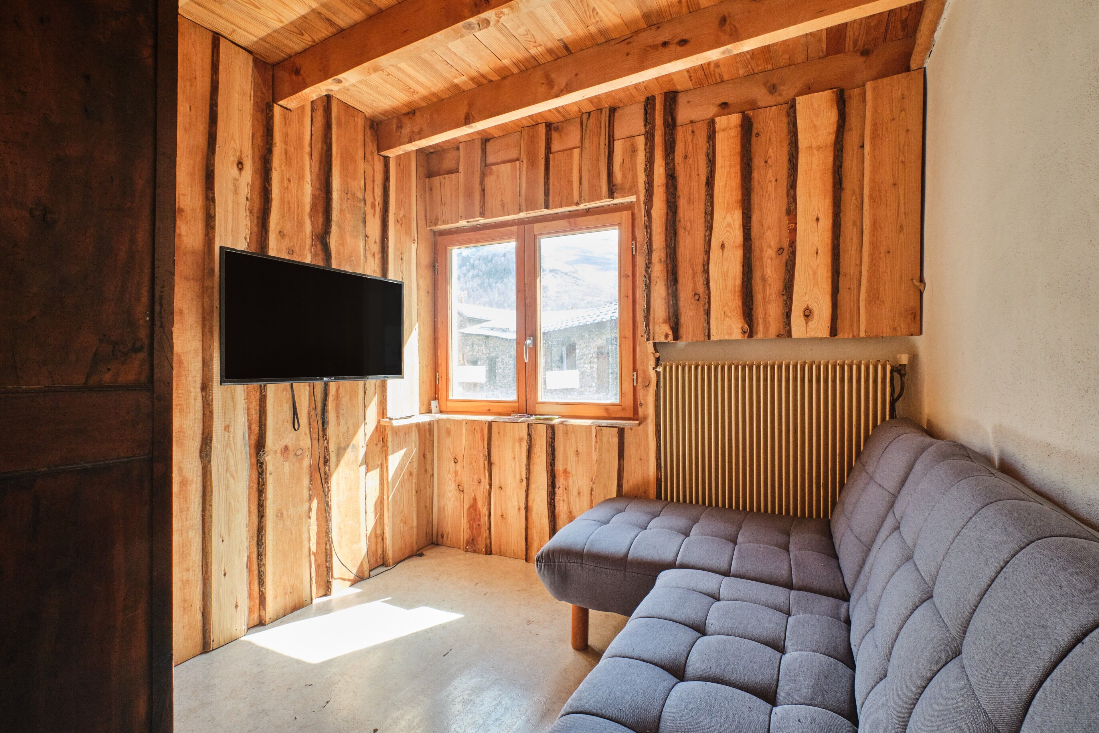 A wooden interior with a flat-screen TV mounted on the wall, a window, and a sofa.