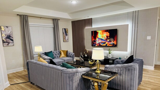 Ein modernes Wohnzimmer mit grauen Polster-Sofas, einem schwarzen Couchtisch mit goldenen Beinen und einem Flachbildfernseher, der ein Feuer zeigt.