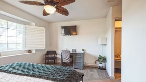A bedroom with a bed, a desk, a chair, a ceiling fan, a TV, and a window.