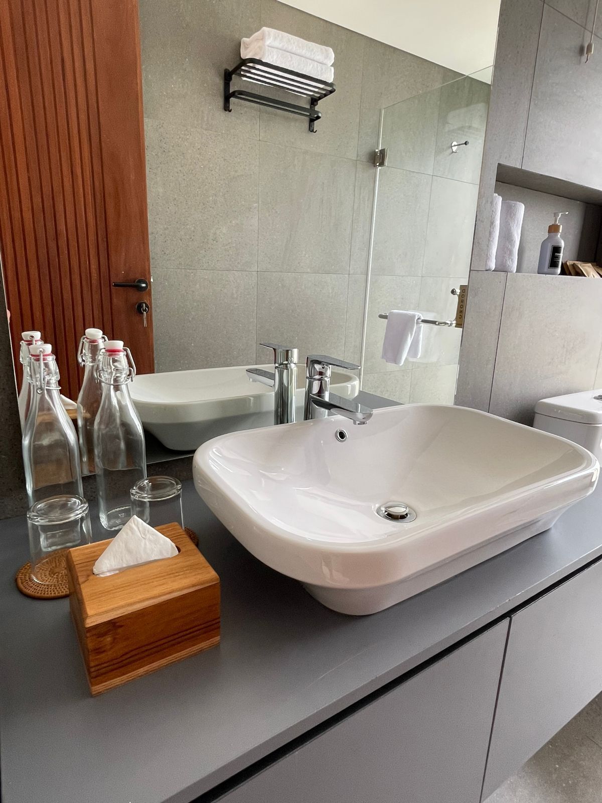 A modern bathroom with a large white sink, a wooden side table with glass bottles and a tissue box, a towel rack with two towels, and a mirror reflecting the shower area.