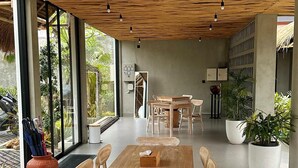 A spacious dining area with a wooden table and chairs, a ceiling with a wavy design, and large windows allowing natural light. - Widji Garden Villa (Pecatu)