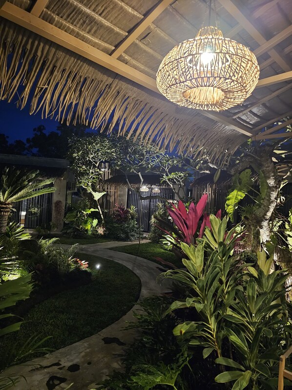 A well-lit outdoor area with a thatched roof, a hanging light fixture, and a variety of tropical plants. - Widji Garden Villa (Pecatu)