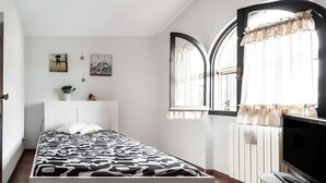 Un dormitorio con cama, televisor, radiador y ventana con cortinas.