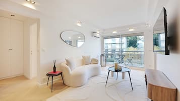 A modern living room with a white sofa, a round mirror, a small table with candles, and a large window offering a view of the outside.