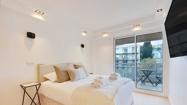 A modern bedroom with a large bed, a small side table, and a balcony with a table and chairs. - Charming Apt with AC & balcony (Suresnes)