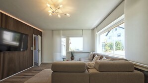 Un salon moderne avec un canapé d’angle, un téléviseur à écran plat fixé sur un panneau en bois et un luminaire suspendu au plafond.