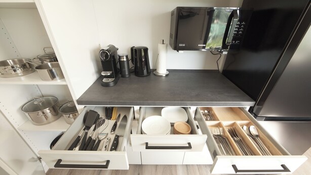 Une cuisine moderne avec un réfrigérateur noir, un four à micro-ondes et une machine à café posés sur le plan de travail. On y trouve des tiroirs blancs contenant des ustensiles et de la vaisselle.