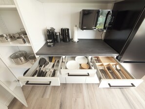 Een moderne keuken met een zwarte koelkast, een magnetron en een koffiezetapparaat op het aanrecht. Er zijn witte lades met bestek en serviesgoed erin.