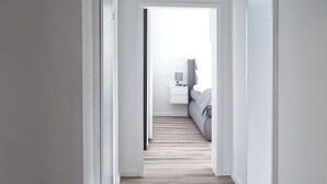 Un couloir au plancher de bois menant à une chambre à coucher équipée d’un lit et d’une table de chevet.