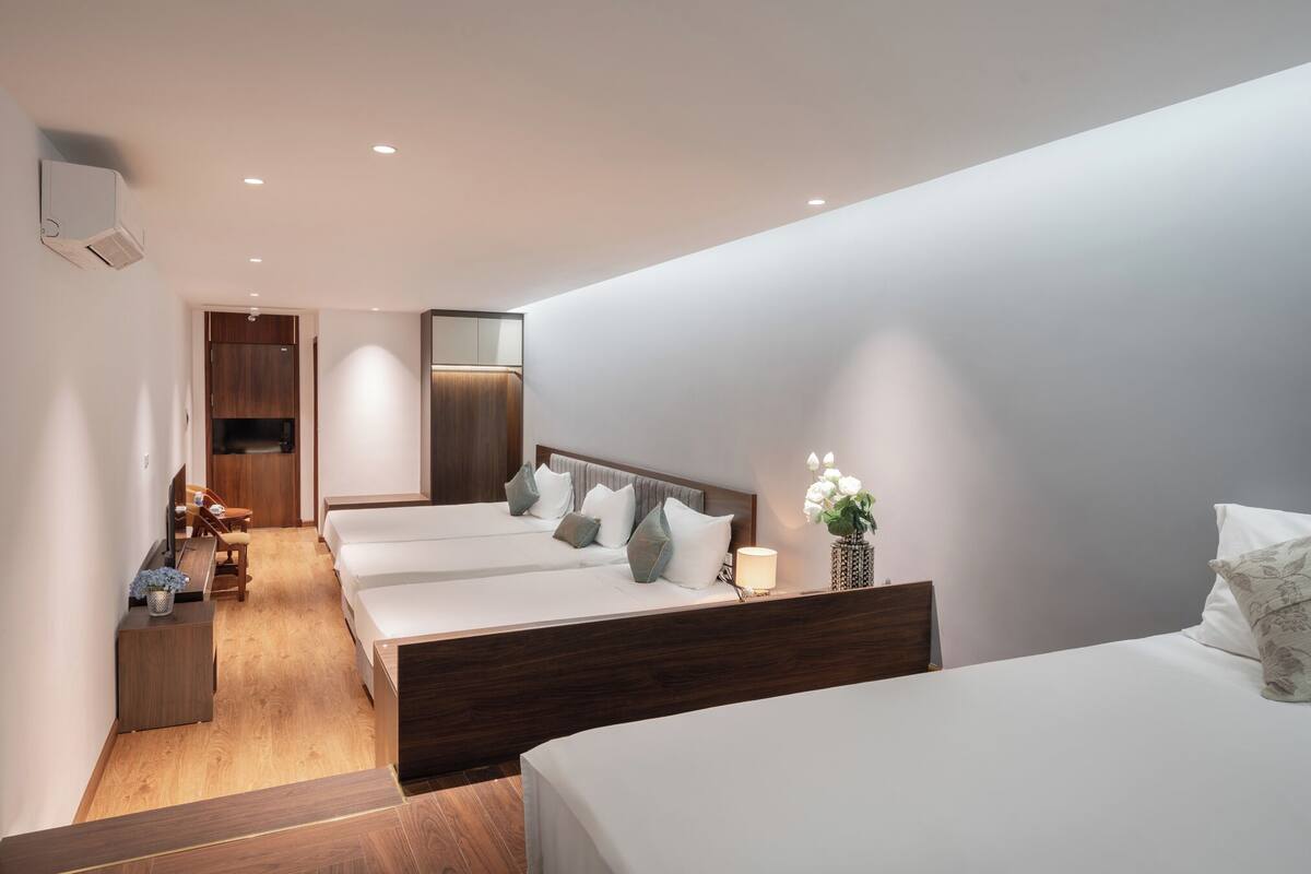 A modern hotel room with a wooden floor, a bed with a wooden headboard, a desk, a chair, and a small table with a plant.