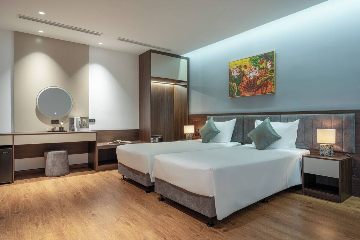 A hotel room with two beds, a desk, a mirror, a nightstand with a lamp, and a painting on the wall.