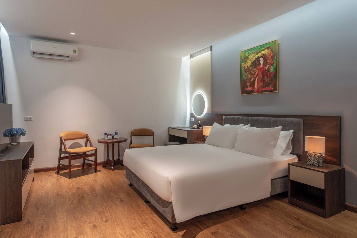 A hotel room with a bed, bedside tables, a desk, chairs, a mirror, and a painting on the wall.