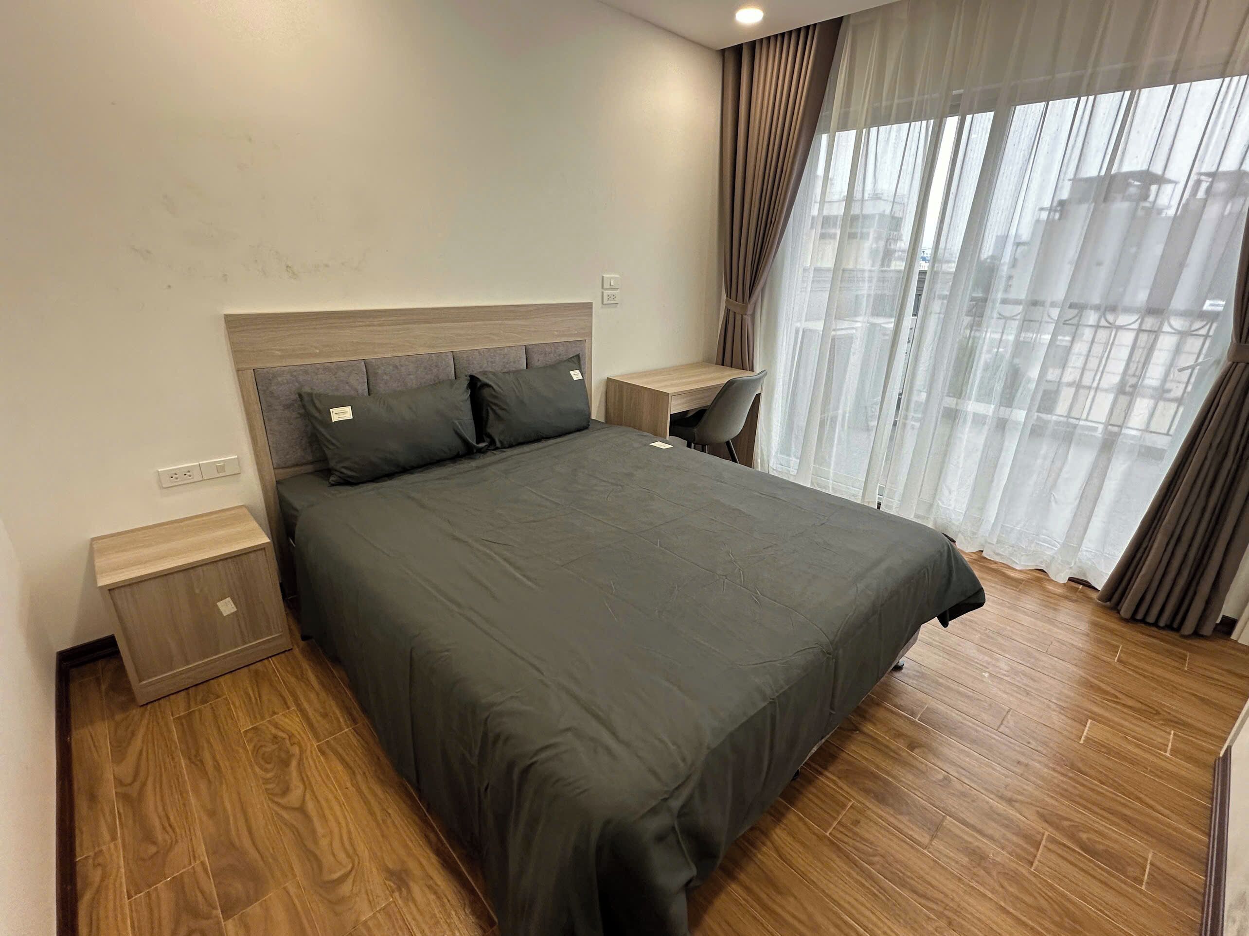 A modern hotel room with a large bed, wooden flooring, a desk, and a view through sheer curtains.