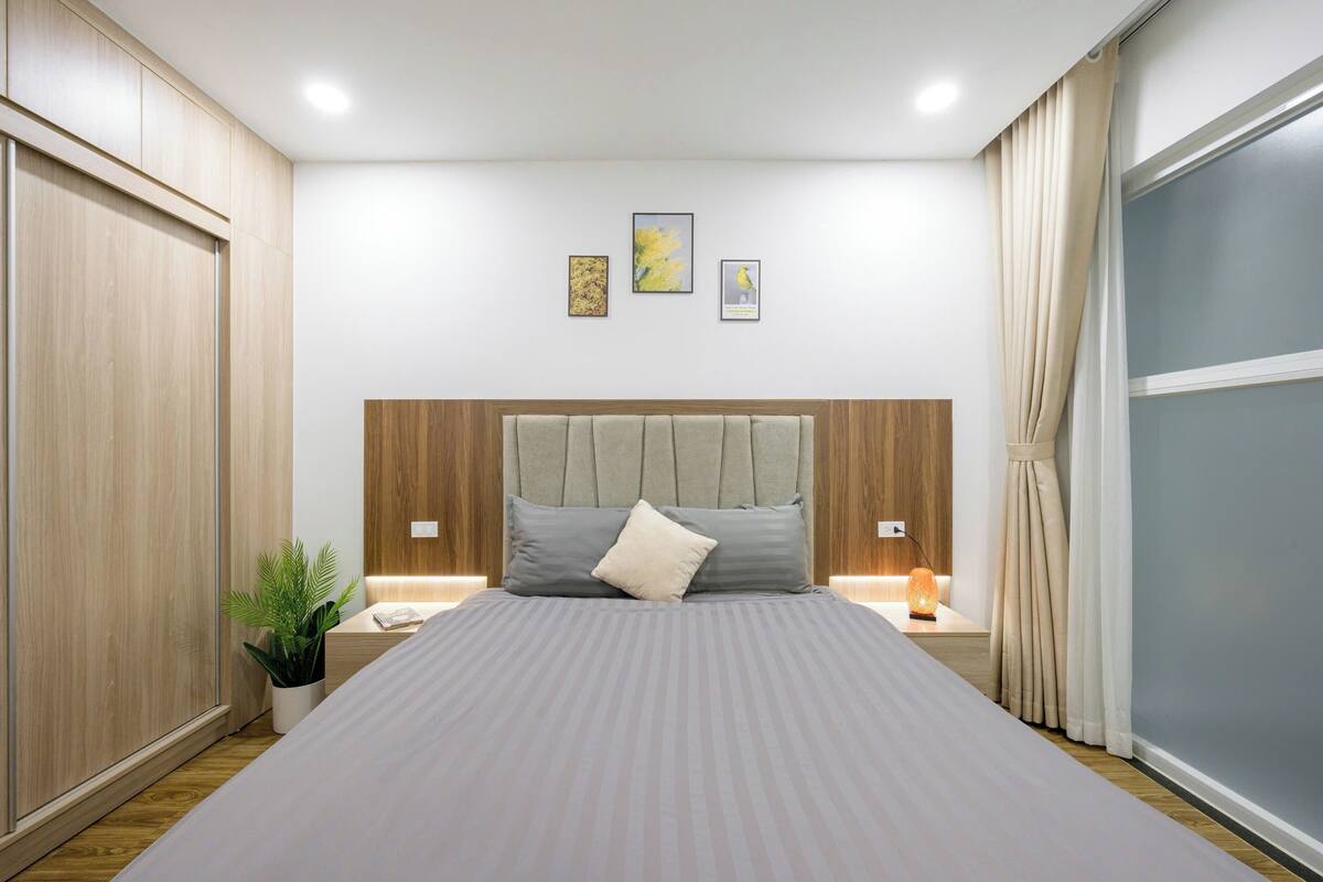 A bedroom with a bed, wooden headboard, bedside tables, a nightstand, a potted plant, and a window with curtains.