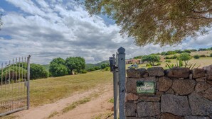 A stone wall with a sign, a metal gate, and a dirt path leading to a green landscape with trees and a building in the background.