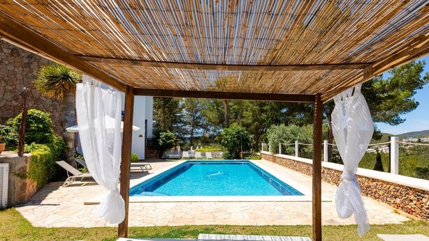 Uma área de piscina com pérgola, piso de pedra e cortinas brancas.