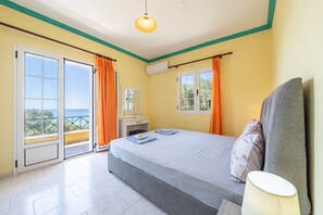A bedroom with a bed, a nightstand, a lamp, and a view of the sea through large windows.