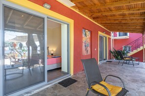 Un espace extérieur abrité, doté d’une porte-fenêtre coulissante donnant sur une pièce, d’une chaise avec un coussin jaune et d’un escalier avec un mur rouge et une rampe noire.