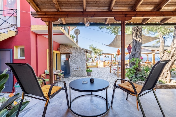 Een overdekt terras met twee stoelen en een tafel, een stenen muur en een rood gebouw op de achtergrond.