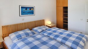 Ein Doppelbett mit blau-weiß karierter Bettwäsche, ein Nachttisch mit Lampe, ein Holz-Kopfstück und ein Kleiderschrank mit offenen Regalen.