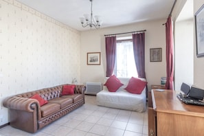 Et værelse med en brun lædersofa, en hvid seng, et træskrivebord med en computer, en radiator og et vindue med gardiner.