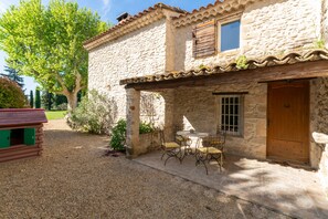 A stone building with a wooden door, a window with shutters, and a patio with a table and chairs.