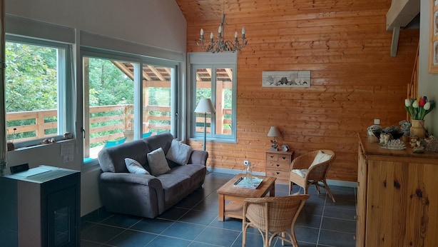 A cozy room with a wooden ceiling, a sofa, a coffee table, and a chair. There are windows with a view of greenery outside.