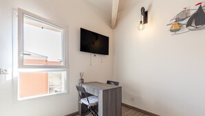 A small room with a TV mounted on the wall, a foldable chair, a small table, and a window with a view. - Bright, Newly Renovated Apartment with Wi-Fi and Air Conditioning (Sanary-sur-Mer)