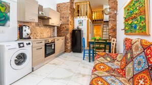 A compact kitchen with a washing machine, a dining area with a colorful sofa, and a brick wall archway.