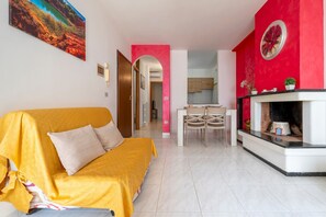 Un salon avec un canapé jaune, un coin repas avec une table et des chaises, un espace cuisine et une cheminée.
