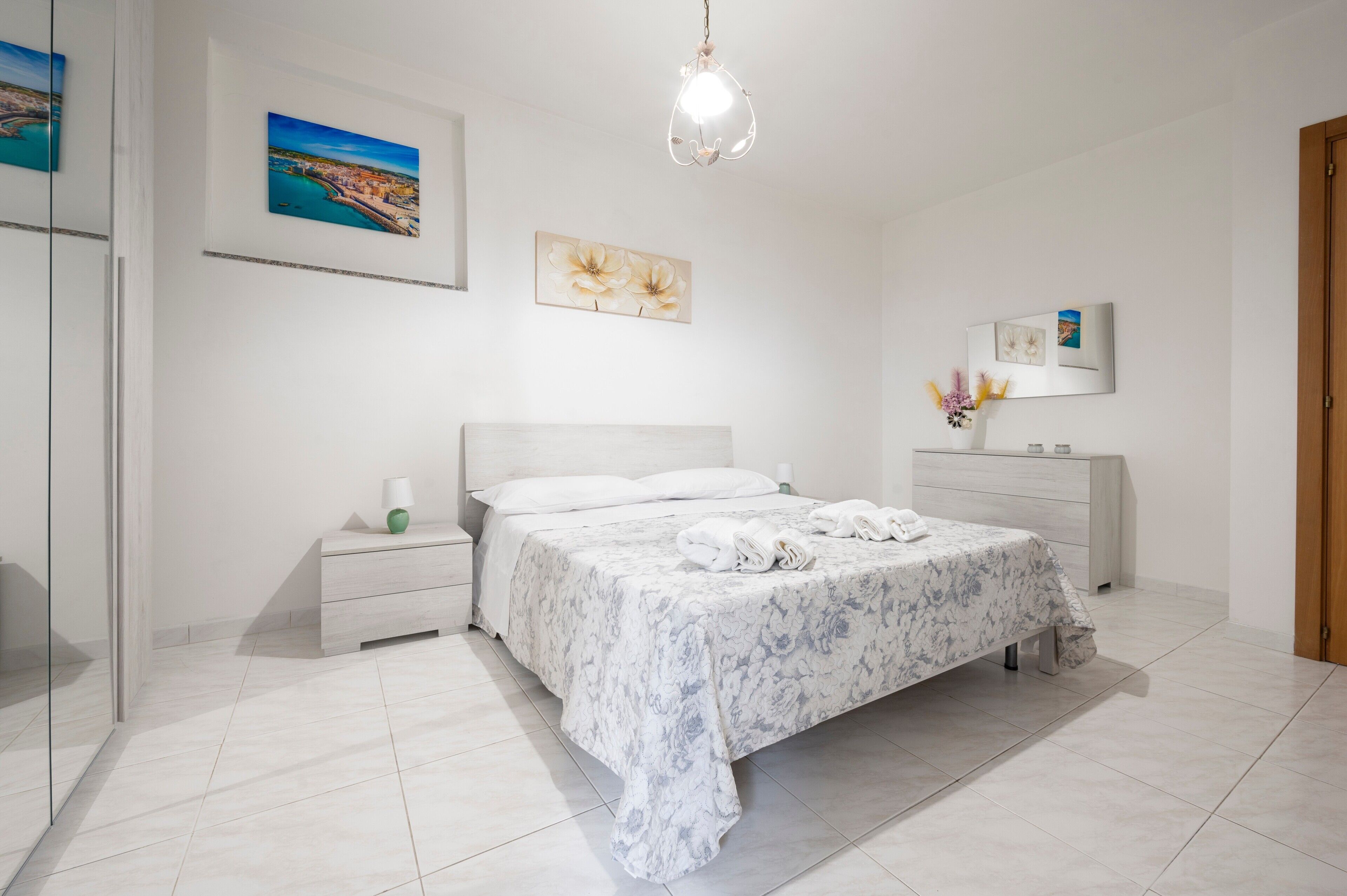 A bedroom with a bed, bedside tables, a nightstand, a mirror, and artwork on the walls.