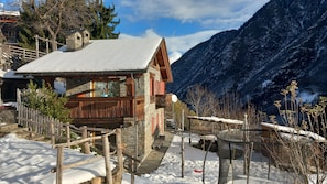 A wooden chalet with a stone foundation, a wooden deck, and a view of snow-covered mountains.