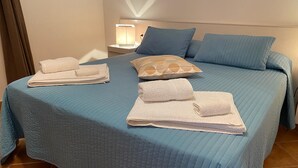 Cama bem arrumada com roupa de cama azul, almofada com padrão geométrico e duas toalhas brancas dobradas.