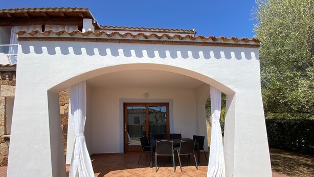 Uma varanda coberta com mesa e cadeiras, uma porta que dá para uma entrada coberta, e um telhado de azulejos.