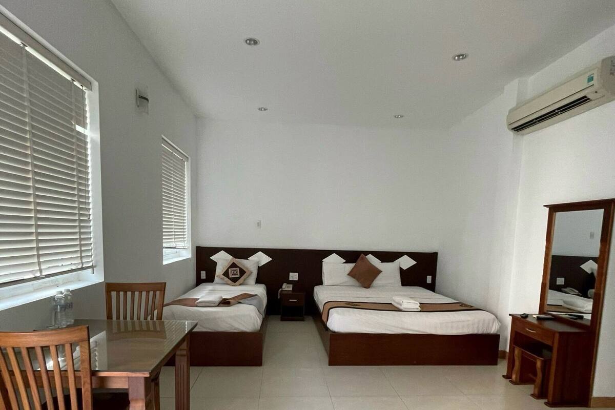 A hotel room with two beds, a wooden table, a chair, a small bedside table, and a mirror.