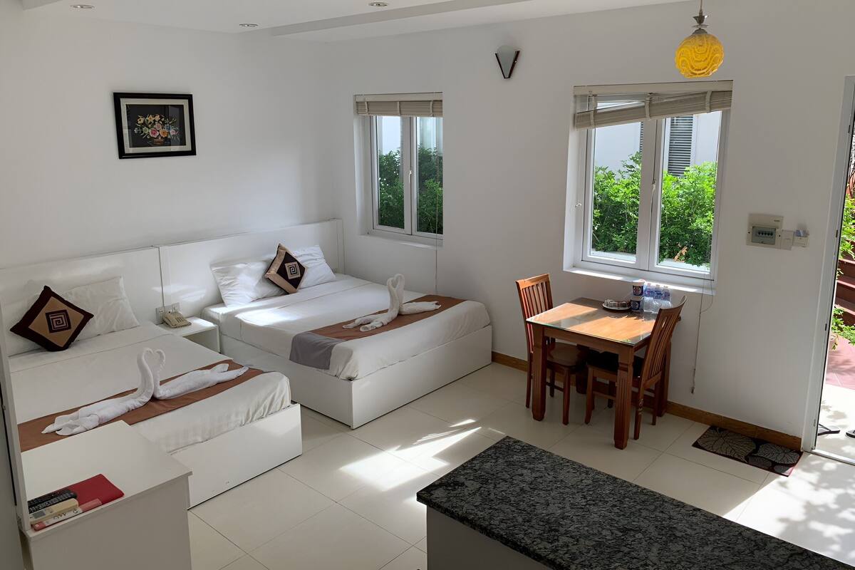 A modern, well-lit room with a bed, a small table, and a chair. There are windows allowing natural light and a view of greenery outside.
