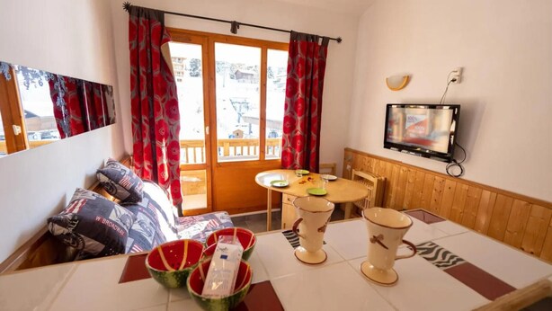 A room with a sofa, a dining table with a pitcher and cups, a television, and a window with curtains. - Mezzanine Studio 'Le Thabor' with Mountain View and Balcony (Modane)