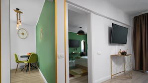 A modern living room with a green accent wall, a dining area with a table and chairs, a television mounted on the wall, and a small side table with decorative items. - Amazon Apartment - Cismigiu Gardens for 2 guests (București)