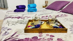 Ein ordentlich bezogenes Bett mit einer blumigen Tagesdecke, ein Tablett mit zwei Gläsern Wasser und Taschentüchern sowie Handtücher in Lila- und Blautönen.
