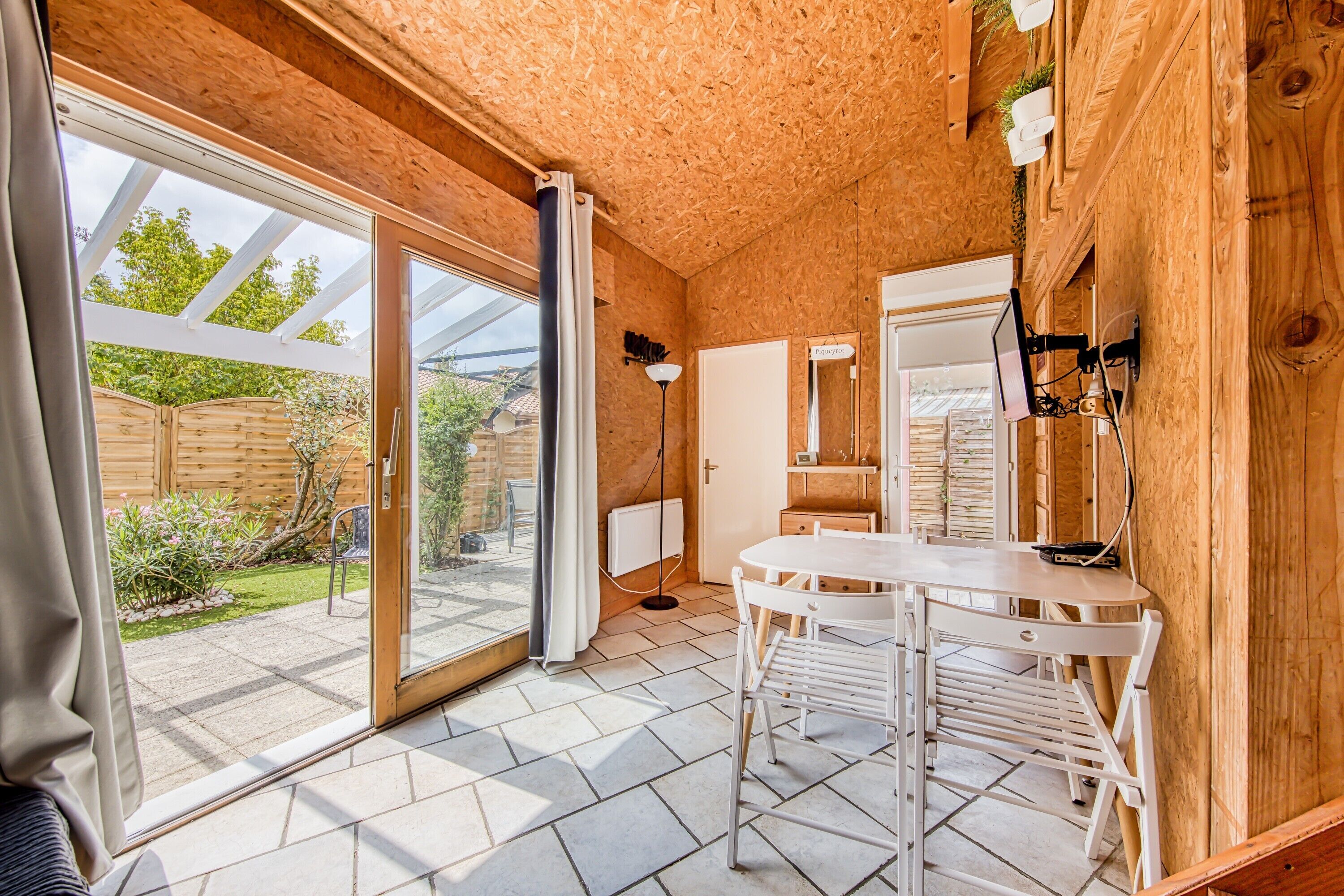 Een houten interieur met een schuifglasdeur naar een tuin, een witte tafel met stoelen en een spiegel aan de muur.